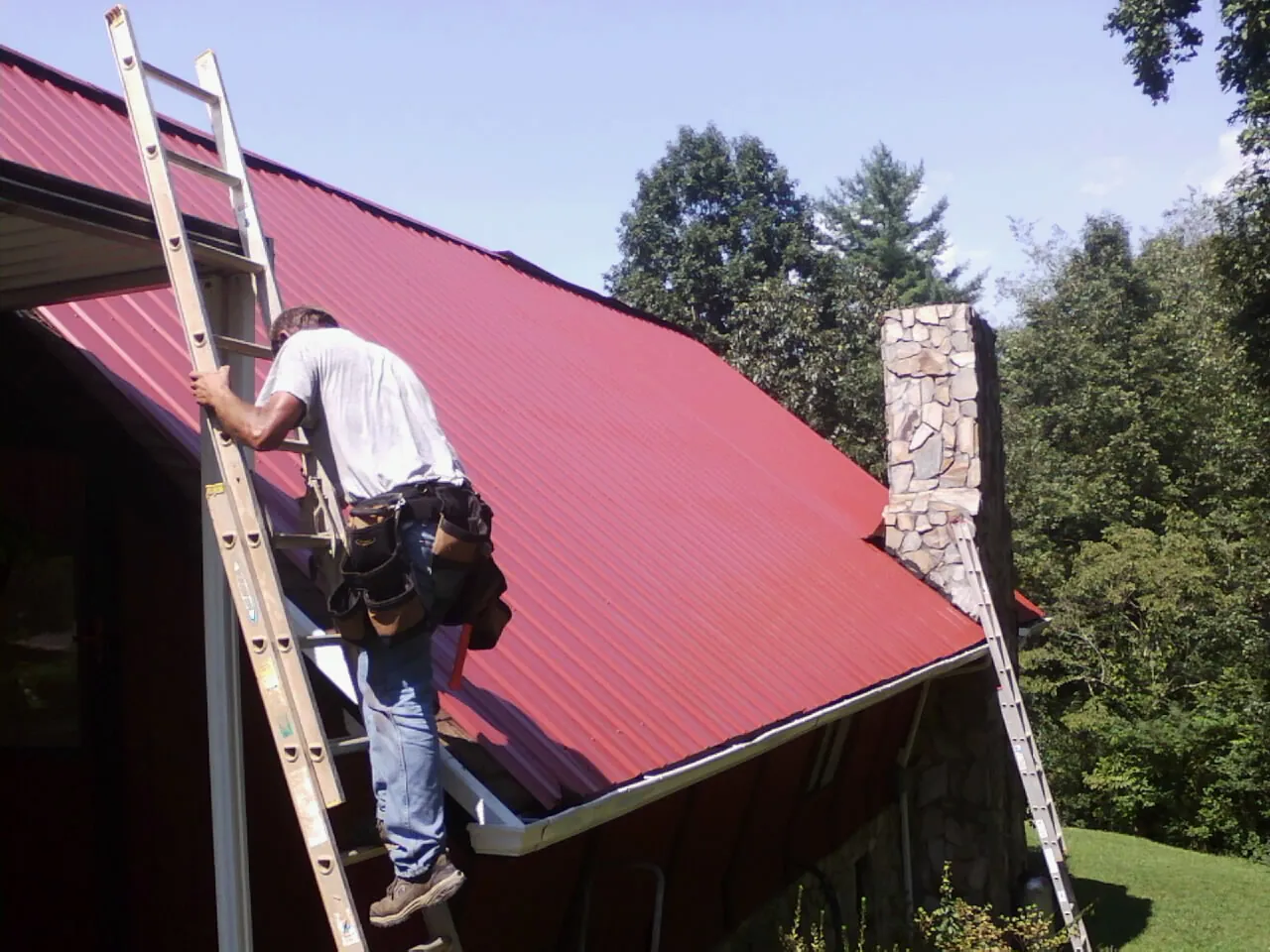Roofer on a ladder beside a red metal roof and stone chimney — Soffit & Fascia Repair by Roofing Brothers in Frazeysburg
